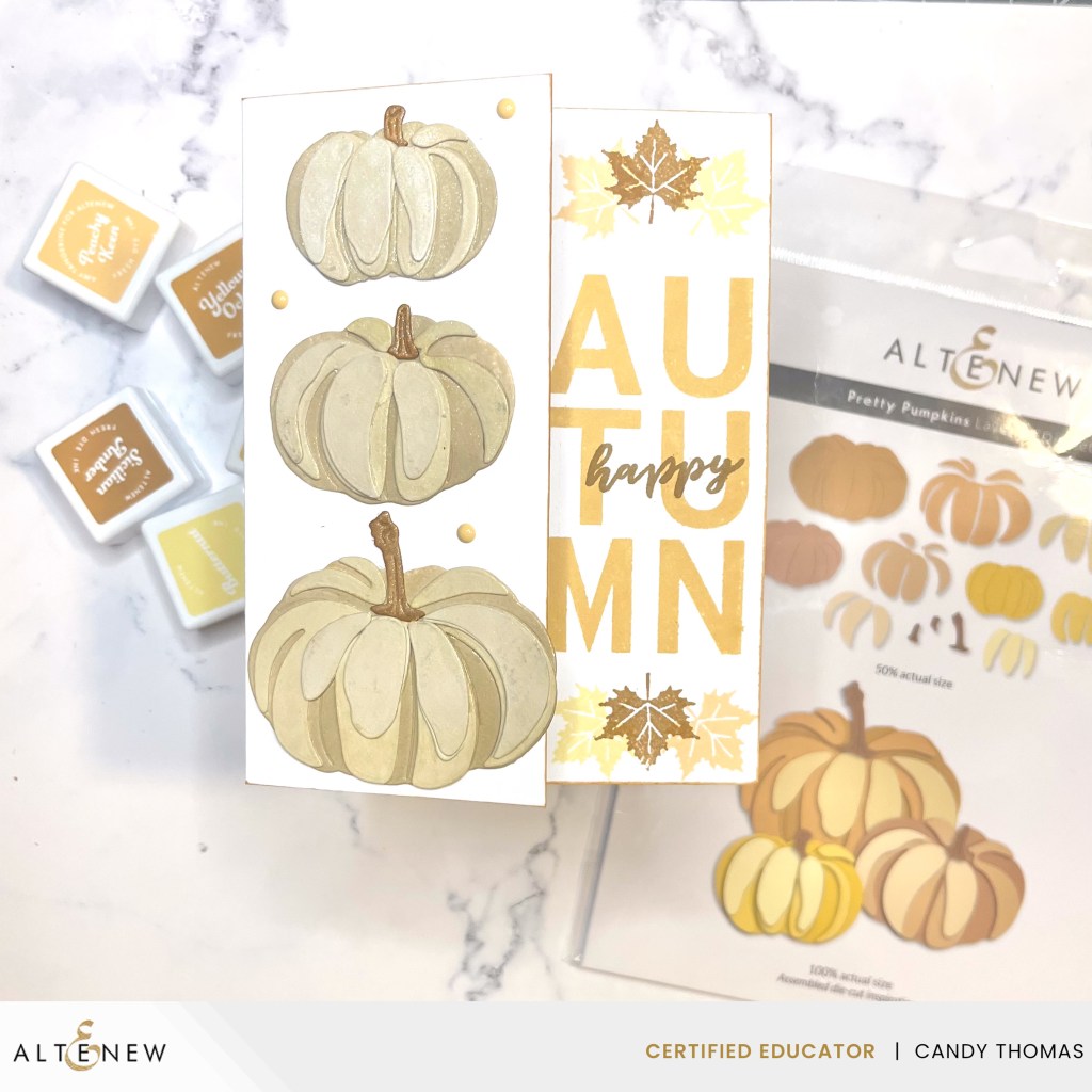 A handmade card featuring three illustrated white pumpkins, the word 'AUTUMN' in golden letters, and autumn leaves, alongside crafting supplies in the background.