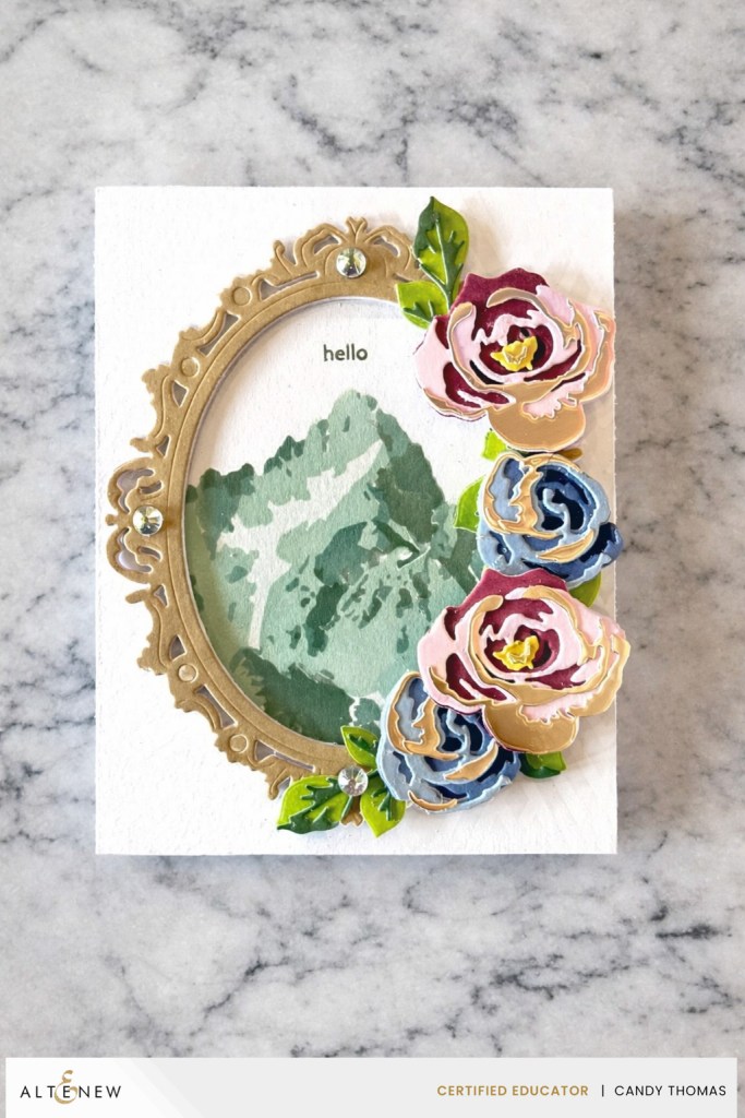 A decorative card featuring a gold ornate frame with the word 'hello,' an artistic depiction of mountains in the background, and vibrant paper flowers in pink and blue on the right side.